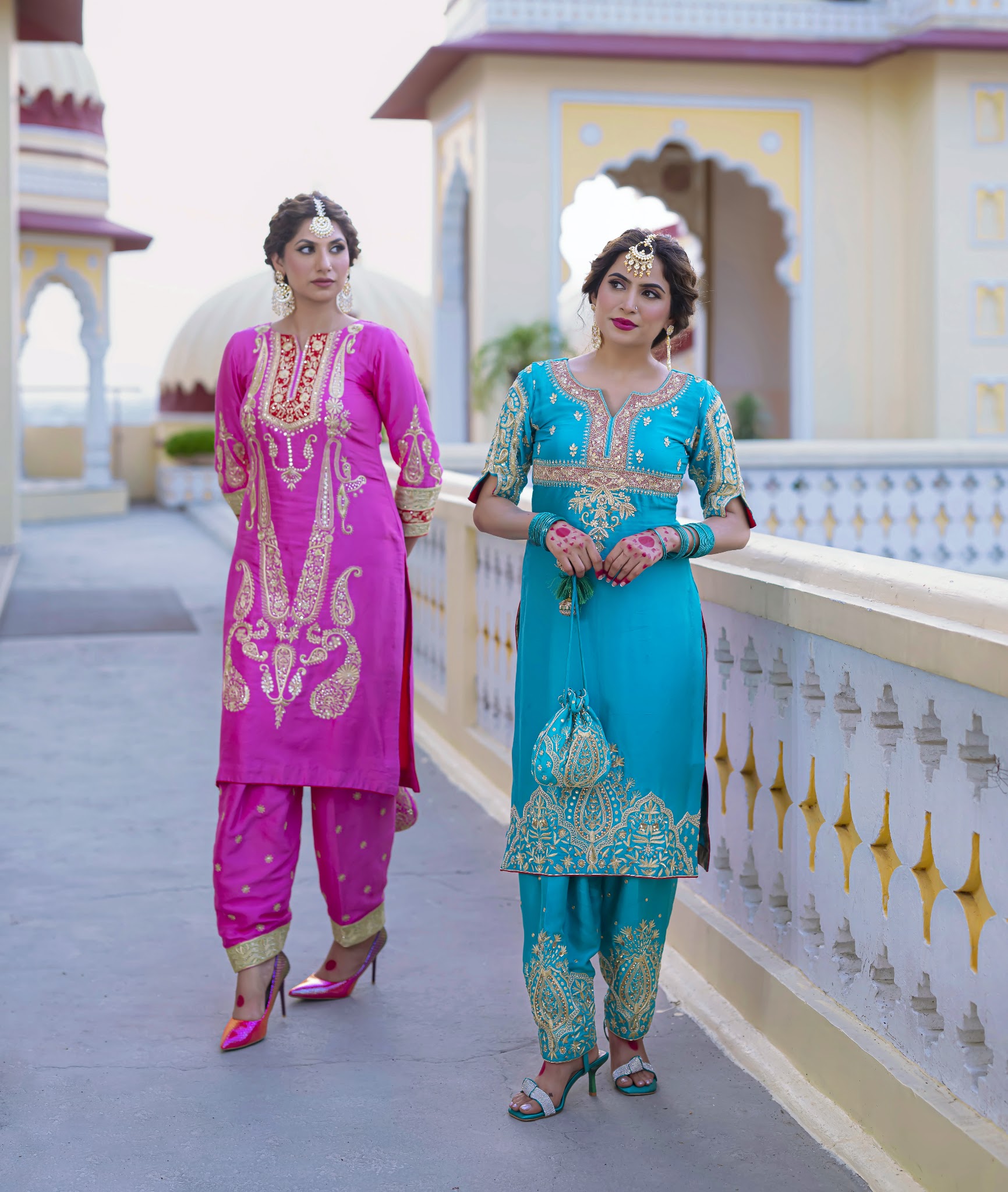 Two women wearing embroidered pink and teal outfits in an ornate courtyard